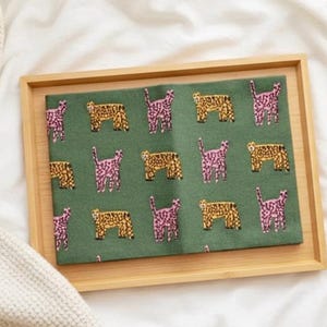 May include: A green rectangular fabric with a repeating pattern of pink and gold animal figures, set inside a light brown wooden tray. The tray is on a white surface, with a white knitted blanket in the background.