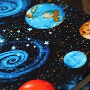 May include: A black fabric with a space theme. The fabric features a blue and white spiral galaxy, blue planets, a brown planet, and white stars on a black background.