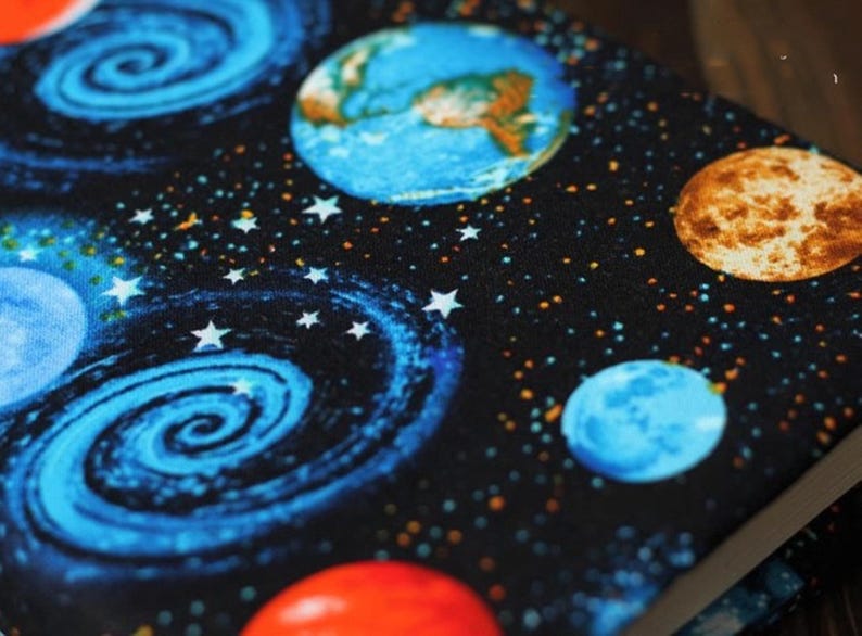 May include: A black fabric with a space theme. The fabric features a blue and white spiral galaxy, blue planets, a brown planet, and white stars on a black background.