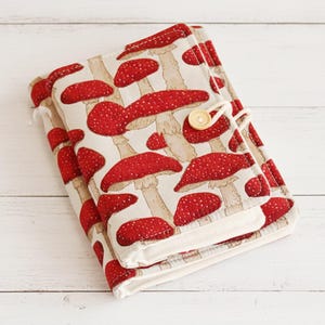 May include: A fabric journal cover featuring a repeating pattern of red and white spotted mushrooms on a beige background. The cover is secured with a button and loop closure. The journal is made of fabric.