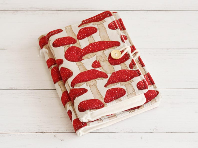 May include: A fabric journal cover featuring a repeating pattern of red and white spotted mushrooms on a beige background. The cover is secured with a button and loop closure. The journal is made of fabric.