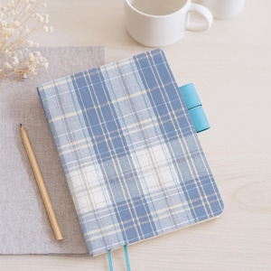 May include: A blue and white plaid journal with a light blue leather closure and two matching ribbon bookmarks. A wooden pencil rests on a gray fabric, next to a white mug and a small potted plant.