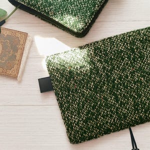 Può includere: Due custodie in tessuto tweed verde con un'etichetta in pelle nera e un nastro nero. Le custodie sono rettangolari e hanno un aspetto strutturato con macchie bianche e rosse. Le custodie sono su una superficie di legno chiaro.