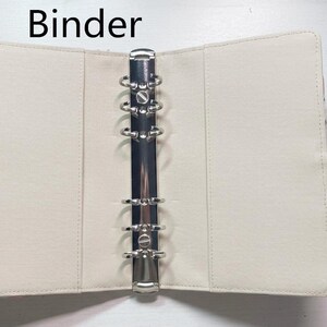 Op de afbeelding: Open, cr&egrave;mekleurige ordner met een zilveren metalen ringmechanisme. De ordner is gemaakt van een glad materiaal en heeft een wit koordje. Het woord "Binder" staat bovenaan.