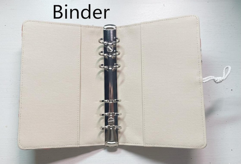 Op de afbeelding: Open, cr&egrave;mekleurige ordner met een zilveren metalen ringmechanisme. De ordner is gemaakt van een glad materiaal en heeft een wit koordje. Het woord "Binder" staat bovenaan.