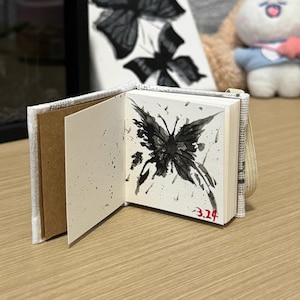 Może przedstawiać: Mały, biały notes w okładce z tkaniny z czarnym rysunkiem motyla na wewnętrznej stronie. Notes ma brązową okładkę z papieru i czarny pasek z tkaniny. Data "3.24" jest napisana czerwonym atramentem w prawym dolnym rogu strony.
