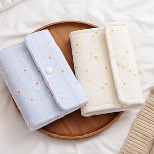Puede incluir: Dos carteras con estampado floral en azul claro y crema, expuestas sobre una bandeja de madera. La cartera azul claro presenta bordados de margaritas blancas, mientras que la cartera crema tiene detalles de margaritas amarillas. Cada cartera tiene un cierre a presión.