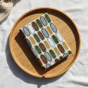 Pode incluir: Um artigo retangular de tecido com um padr&atilde;o oval em branco, azul, castanho, verde e amarelo &eacute; exibido em uma bandeja redonda de madeira. A bandeja est&aacute; sobre uma superf&iacute;cie branca, com uma por&ccedil;&atilde;o de um artigo de malha creme vis&iacute;vel.