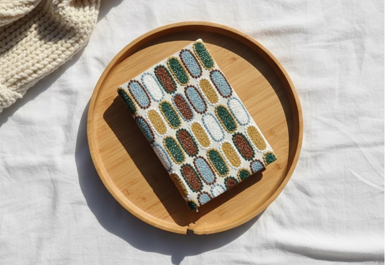 Pode incluir: Um artigo retangular de tecido com um padr&atilde;o oval em branco, azul, castanho, verde e amarelo &eacute; exibido em uma bandeja redonda de madeira. A bandeja est&aacute; sobre uma superf&iacute;cie branca, com uma por&ccedil;&atilde;o de um artigo de malha creme vis&iacute;vel.