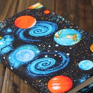 May include: A black book cover with a space-themed design. The cover features planets, stars, and swirling galaxies in shades of blue, orange, and yellow.