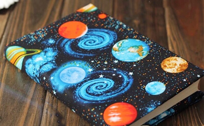 May include: A black book cover with a space-themed design. The cover features planets, stars, and swirling galaxies in shades of blue, orange, and yellow.
