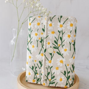 Puede incluir: Cartera bordada beige con margaritas blancas con centros amarillos y tallos verdes. La cartera tiene un cierre de botón y se muestra en una bandeja de madera. Un jarrón de cristal con flores blancas está en el fondo.