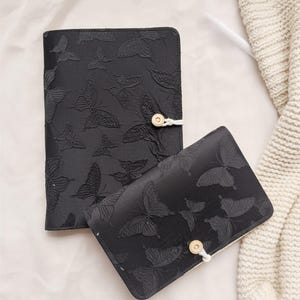 Può includere: Due quaderni neri con un motivo a farfalle. Il quaderno più grande è chiuso con un bottone e uno spago. Il quaderno più piccolo è aperto. Entrambi i quaderni presentano un design a farfalla strutturato.
