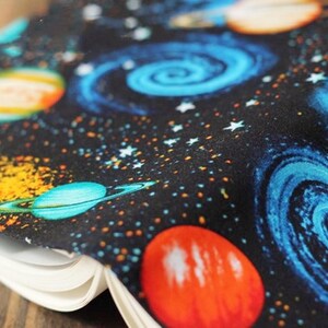 May include: A black fabric with a space-themed print featuring planets, stars, and swirling galaxies in shades of blue, orange, and yellow.