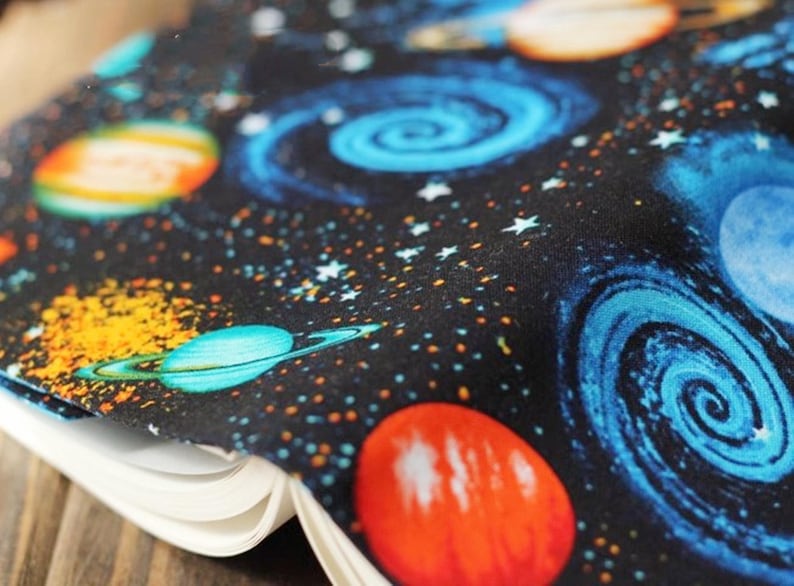 May include: A black fabric with a space-themed print featuring planets, stars, and swirling galaxies in shades of blue, orange, and yellow.