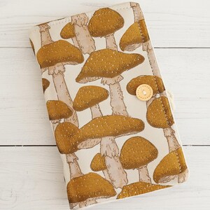 May include: A beige fabric case featuring a repeating pattern of brown mushrooms with white stems. The case has a button closure and a stitched border. The mushroom design is detailed with speckled caps.