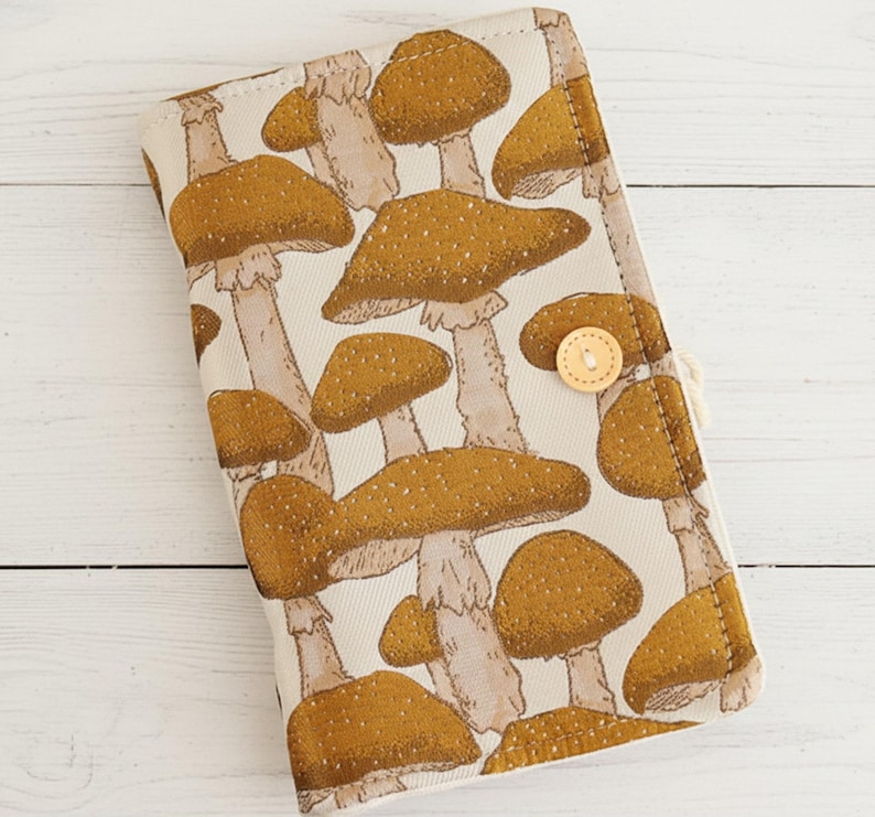 May include: A beige fabric case featuring a repeating pattern of brown mushrooms with white stems. The case has a button closure and a stitched border. The mushroom design is detailed with speckled caps.