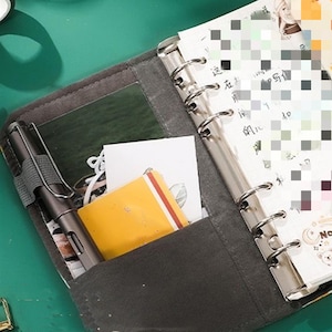 May include: A gray fabric bound planner with a silver ring binder. The planner has a clear plastic pocket on the front cover, which holds a yellow notepad and a pen. The planner is open to a page with a calendar.