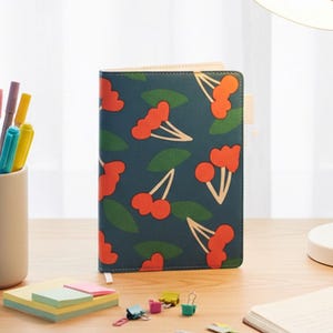 Peut inclure: Un cahier bleu canard avec un motif de cerises, un pot à crayons avec des stylos et des ciseaux, et des post-it sont posés sur un bureau en bois. Une lampe de bureau est à l'arrière-plan. Le cahier a un signet blanc.