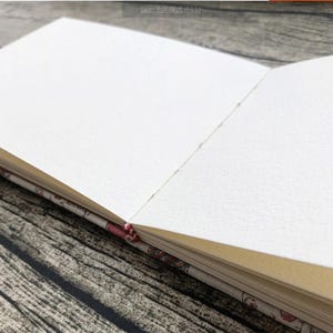 Może przedstawiać: Zbliżenie na pusty, biały notes z kremową okładką. Notes jest otwarty na świeżą, niekreśloną stronę. Okładka jest ozdobiona wzorem kwiatowym.