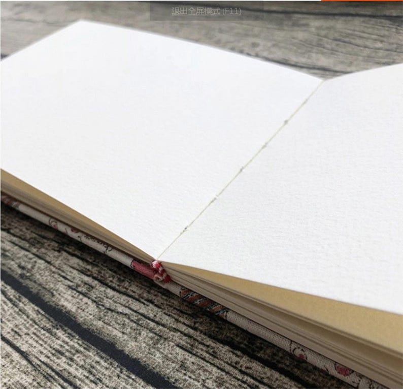 Może przedstawiać: Zbliżenie na pusty, biały notes z kremową okładką. Notes jest otwarty na świeżą, niekreśloną stronę. Okładka jest ozdobiona wzorem kwiatowym.