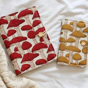 以下が含まれることがあります： キノコの模様の生地ケースが2つ。1つのケースは、クリーム色の背景に白い水玉模様の赤いキノコが特徴です。もう1つのケースは、クリーム色の背景に茶色のキノコがあります。どちらのケースにも木製のボタン留めが付いています。