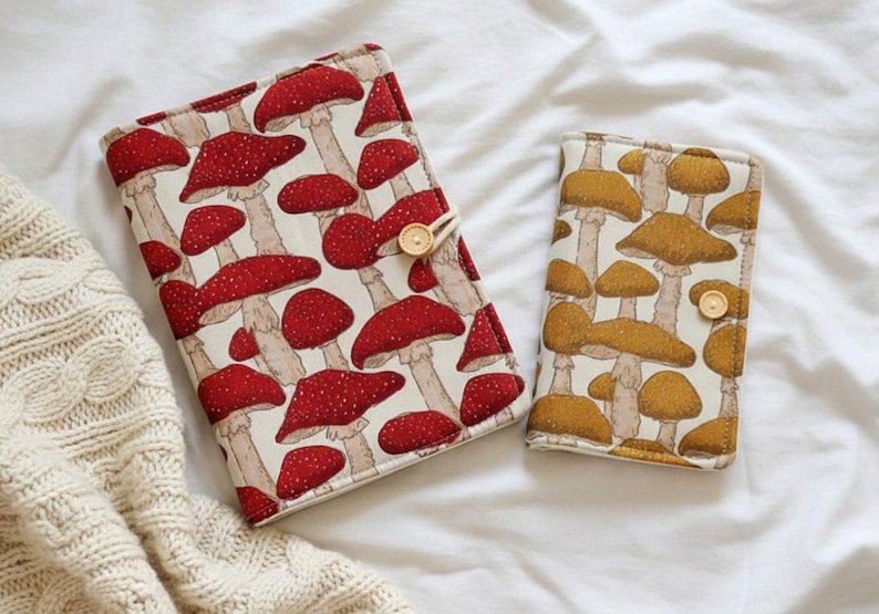 May include: Two fabric cases with mushroom patterns. One case features red mushrooms with white spots on a cream background. The other case has brown mushrooms on a cream background. Both cases have a wooden button closure.