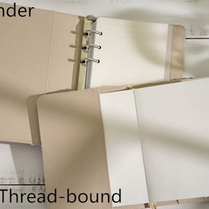 Op de afbeelding: Beige ordner met een zilveren metalen ringmechanisme, open om blanco witte pagina's te onthullen. Het woord "Binder" staat linksboven en "Thread-bound" onderaan. De ordner ligt op een licht houten oppervlak.
