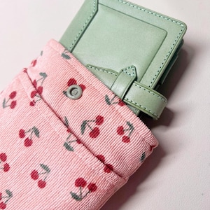 May include: A light green leather wallet with a strap closure is tucked into a pink corduroy pouch with a cherry print.