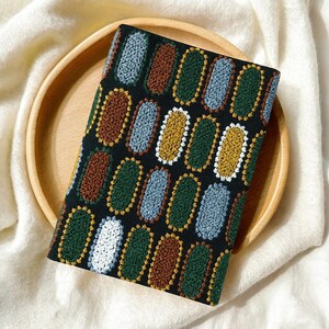 Pode incluir: Um item de tecido preto com um padr&atilde;o repetido de desenhos ovais coloridos em marrom, verde, azul, amarelo e branco. O item est&aacute; sobre uma bandeja redonda de madeira, colocada sobre uma superf&iacute;cie branca texturizada.