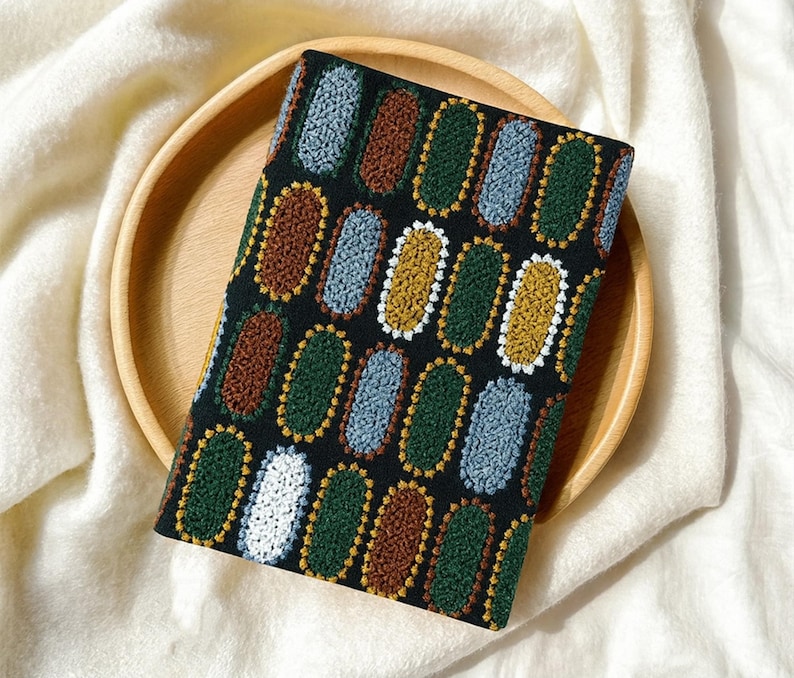 Pode incluir: Um item de tecido preto com um padr&atilde;o repetido de desenhos ovais coloridos em marrom, verde, azul, amarelo e branco. O item est&aacute; sobre uma bandeja redonda de madeira, colocada sobre uma superf&iacute;cie branca texturizada.