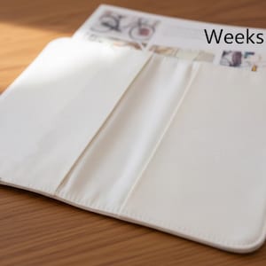 Pode incluir: Uma capa de planejador aberta, de cor creme, com o texto "Weeks inner" vis&iacute;vel em uma p&aacute;gina parcialmente inserida. A capa &eacute; feita de um material liso e repousa sobre uma superf&iacute;cie de madeira.