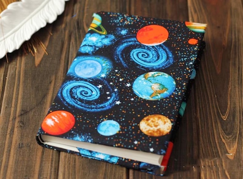 May include: A book cover with a space-themed design. The cover features a black background with blue and orange planets, stars, and swirling galaxies.