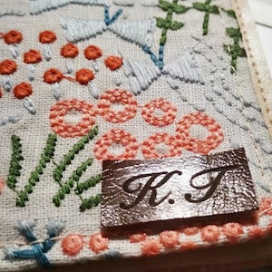 May include: A white fabric with a floral embroidery design in shades of blue, green, and orange. A brown leather tag with the initials "K.T." is sewn onto the fabric.
