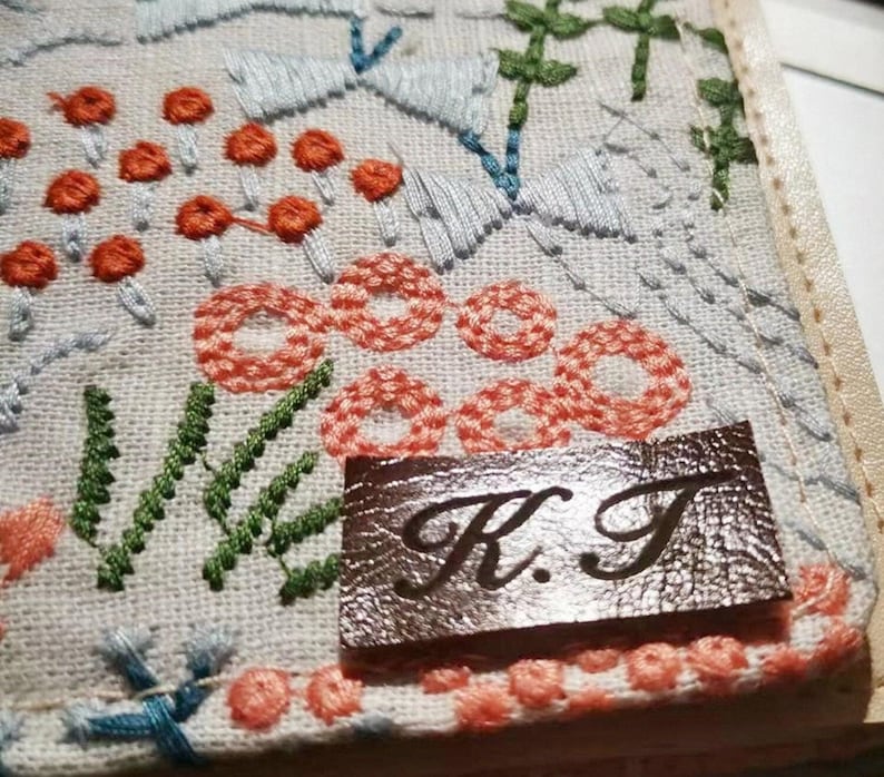 May include: A white fabric with a floral embroidery design in shades of blue, green, and orange. A brown leather tag with the initials "K.T." is sewn onto the fabric.