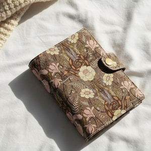 Puede incluir: Una cartera marrón con estampado floral y cierre de botón a presión dorado. La cartera presenta un diseño repetido de flores y follaje en tonos rosa, crema y marrón. La cartera está cerrada y sobre una superficie blanca.
