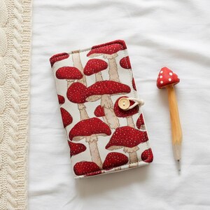 May include: A notebook with a red mushroom pattern and a wooden mushroom-shaped pen. The notebook has a beige background with red mushrooms and a button closure. The pen has a red mushroom top with white spots.