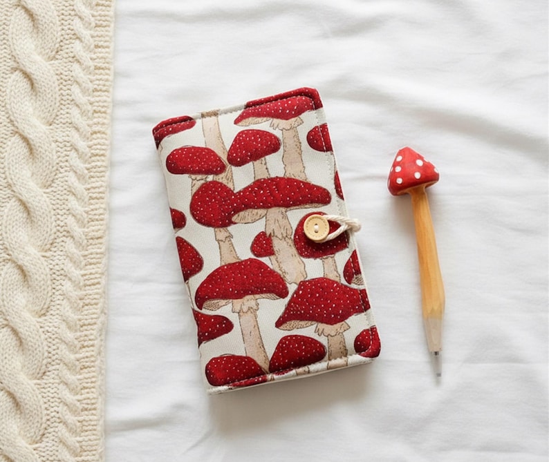 May include: A notebook with a red mushroom pattern and a wooden mushroom-shaped pen. The notebook has a beige background with red mushrooms and a button closure. The pen has a red mushroom top with white spots.