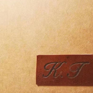 Pode incluir: Uma etiqueta retangular de couro castanho com as iniciais "K.T." gravadas. A etiqueta est&aacute; colocada sobre um fundo texturizado bege. As letras est&atilde;o em escrita cursiva.