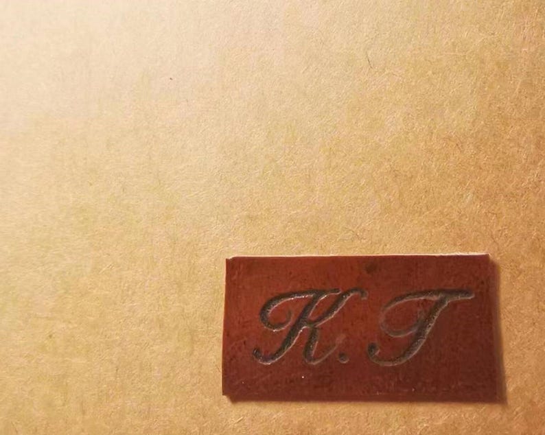 Pode incluir: Uma etiqueta retangular de couro castanho com as iniciais "K.T." gravadas. A etiqueta est&aacute; colocada sobre um fundo texturizado bege. As letras est&atilde;o em escrita cursiva.