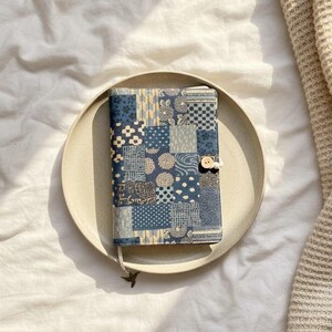 Op de afbeelding: Een blauw-witte patchwork boekomslag met een houten knoopsluiting. De omslag ligt op een beige bord, tegen een witte achtergrond. Het ontwerp bevat verschillende geometrische en bloemmotieven.