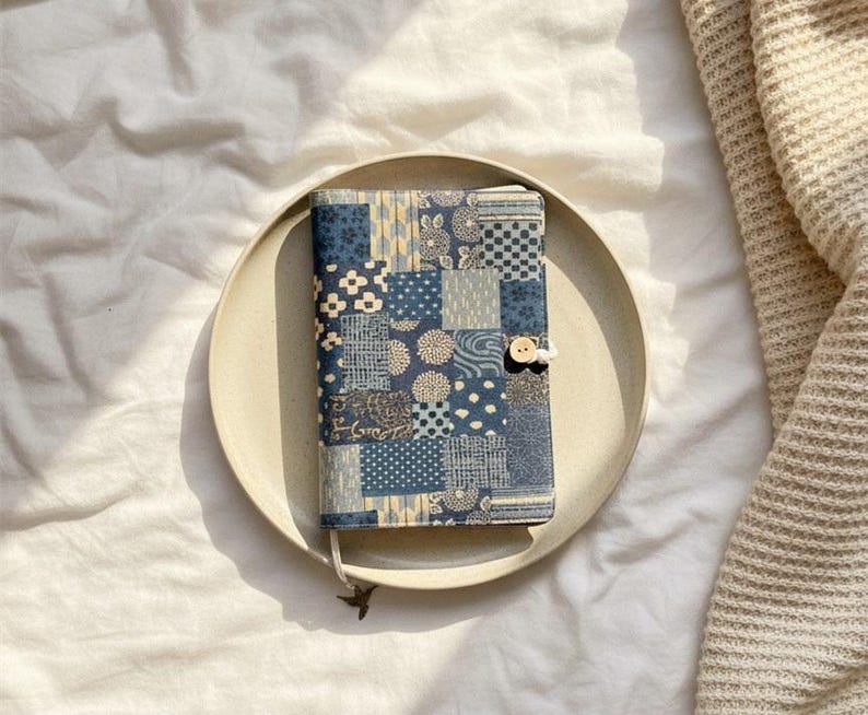 Op de afbeelding: Een blauw-witte patchwork boekomslag met een houten knoopsluiting. De omslag ligt op een beige bord, tegen een witte achtergrond. Het ontwerp bevat verschillende geometrische en bloemmotieven.