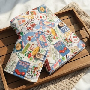Puede incluir: Una funda de diario de tela con un tema de tarta de cerezas y cerezas. La funda es blanca con un collage de gráficos con temas de cerezas, que incluyen texto, cerezas y rebanadas de pastel. Tiene un cierre de botón de madera y descansa sobre una bandeja de madera.