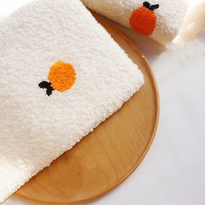 May include: White, fluffy towels with embroidered orange fruit designs. The towels are placed on a wooden tray, with one towel folded and another partially visible. The orange fruit embroidery has a black stem and leaf.