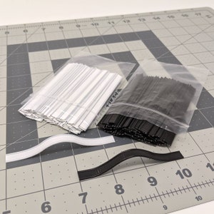 May include: Two bags of white and black plastic-coated metal ties. The ties are in separate bags and are arranged in a grid pattern. The bags are on a gray cutting mat with white markings.