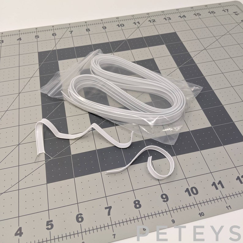 May include: A white plastic bag containing a large quantity of white, flexible, plastic nose wire for face masks. The nose wire is coiled and arranged in a circular shape. The bag is sitting on a gray gridded surface.