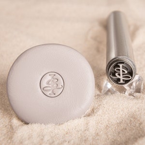 May include: A close-up of a silver metal stamp with a monogram design. The stamp is on a white surface with sand.