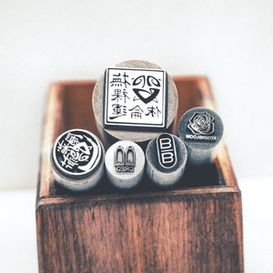 Può includere: Un set di sei timbri in legno con diversi disegni. I timbri sono in una scatola di legno. I timbri presentano disegni come un fantasma, un fiore e un logo con caratteri giapponesi.