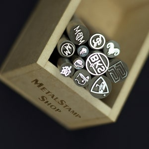 Puede incluir: Una caja de madera llena de sellos de metal, cada uno con un diseño diferente. La caja está etiquetada "METAL STAMP SHOP".