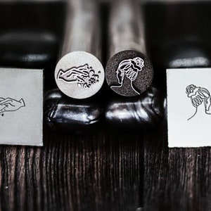 Può includere: Tre teste di timbro in metallo con diversi disegni. La prima testa di timbro ha un disegno di una mano con stelle e lune. La seconda testa di timbro ha un disegno di un viso di donna con i capelli raccolti in uno chignon. La terza testa di timbro ha un disegno di una testa di donna con i capelli raccolti in uno chignon.